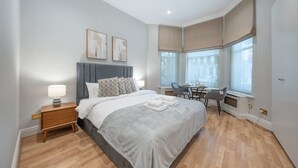 Een slaapkamer met een bed, nachtkastje, lamp en uitzicht op de stad door grote ramen.