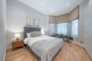 Ein Schlafzimmer mit einem Bett, einem Nachttisch, einer Lampe und Blick auf die Stadt durch große Fenster.