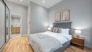 Een moderne slaapkamer met een grijs hoofdeinde, een bed met witte lakens, twee bedlampen en een houten nachtkastje.
