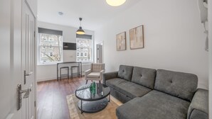 Ein modernes Wohnzimmer mit einem grauen Sofa, einem Glastisch und einem Sessel. Es gibt zwei Pendelleuchten, ein weißes Telefon und gerahmte Kunstwerke an der Wand.
