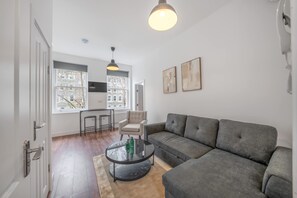Ein modernes Wohnzimmer mit einem grauen Sofa, einem Glastisch und einem Sessel. Es gibt zwei Pendelleuchten, ein weißes Telefon und gerahmte Kunstwerke an der Wand.