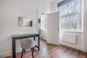 Ein Zimmer mit Holzboden, einem Schreibtisch mit Stuhl, einer weißen Tür und einem Fenster mit Blick auf Gebäude.