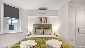 A hotel room with a bed, two bedside lamps, a window with a blind, an air conditioning unit, and a door. - Standard Studio in Knightsbridge (London)
