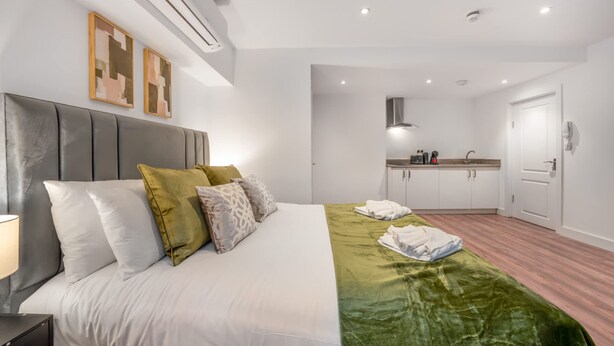 A modern hotel room with a bed, a bedside table, a green throw blanket, a white headboard, and a kitchenette in the background. - Standard Studio in Knightsbridge (London)
