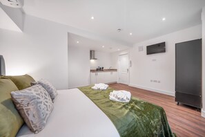 Ein Hotelzimmer mit Bett, grüner Bettdecke, Kissen, einem Fernseher, einem Kleiderschrank und einer Küchenzeile im Hintergrund.