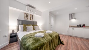 A modern bedroom with a large bed, a bedside table, an air conditioning unit, a wall-mounted light, and a kitchenette area with a stove and coffee maker. - Standard Studio in Knightsbridge (London)