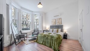 A modern hotel room with a large bed, a seating area with a chair and table, a flat-screen TV, and large windows offering a city view. - Deluxe Ground Floor Studio in Knightsbridge (London)