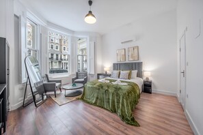 A modern hotel room with a large bed, a seating area with a chair and table, a flat-screen TV, and large windows offering a city view. - Deluxe Ground Floor Studio in Knightsbridge (London)