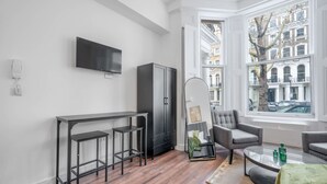 A modern living room with a bar area, a flat-screen TV mounted on the wall, and a large window offering a view of the city. - Deluxe Ground Floor Studio in Knightsbridge (London)