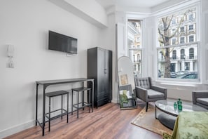 A modern living room with a bar area, a flat-screen TV mounted on the wall, and a large window offering a view of the city. - Deluxe Ground Floor Studio in Knightsbridge (London)
