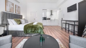 A modern hotel room with a bed, a desk, a chair, a kitchen area, and a TV mounted on the wall. - Deluxe Ground Floor Studio in Knightsbridge (London)