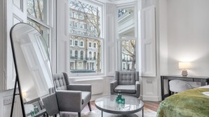 A hotel room with a large window, a round table with a glass top, two grey armchairs, a green bedspread, and a mirror. - Deluxe Ground Floor Studio in Knightsbridge (London)