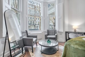 A hotel room with a large window, a round table with a glass top, two grey armchairs, a green bedspread, and a mirror. - Deluxe Ground Floor Studio in Knightsbridge (London)