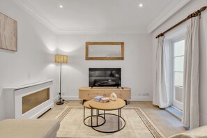 A modern living room with a fireplace, a television, a round coffee table, and a large window with curtains. - Modern Split-level Apartment in Knightsbridge (London)