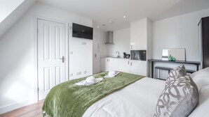 A bedroom with a bed, a green blanket, a pillow, a TV on the wall, and a kitchenette area. - Top Floor Studio With View in Knightsbridge (London)