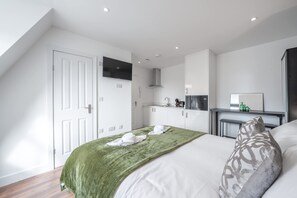 A bedroom with a bed, a green blanket, a pillow, a TV on the wall, and a kitchenette area. - Top Floor Studio With View in Knightsbridge (London)