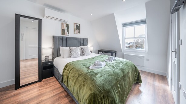 A modern bedroom with a large bed, a bedside table, a nightstand, a mirror, and a window. - Top Floor Studio With View in Knightsbridge (London)