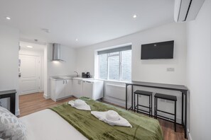 A modern hotel room with a bed, a green throw blanket, two towels, a kitchen area, a window, a flat-screen TV, and a desk with two chairs. - Superior Ground Floor Studio in Knightsbridge (London)