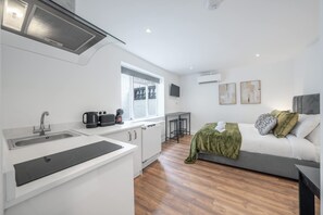 A compact kitchen with a sink, stove, and coffee maker, a small dining area with a table and chairs, a bed with pillows, and a window with a view. - Superior Ground Floor Studio in Knightsbridge (London)