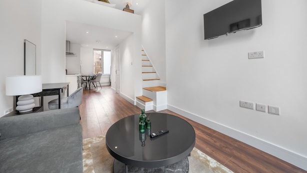 A modern living room with a flat-screen TV mounted on the wall, a round coffee table with remote controls, and a staircase leading to an upper level. - Deluxe Studio Mezzanine With Balcony in Knightsbridge (London)