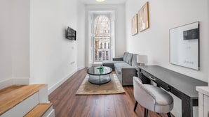 A modern living room with a round glass coffee table, grey sofa, and a dining area with a dark countertop. - Deluxe Studio Mezzanine With Balcony in Knightsbridge (London)