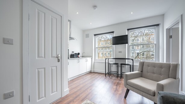 A modern kitchen with white cabinetry, a dining area with a grey sofa, and large windows offering views of buildings. - Charming One Bedroom Flat in Knightsbridge (London)