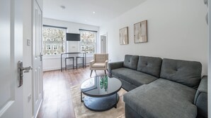 A modern living room with a grey sectional sofa, a round coffee table, and a beige armchair. There are two windows with a view of trees and buildings, and a flat-screen TV mounted on the wall. - Charming One Bedroom Flat in Knightsbridge (London)