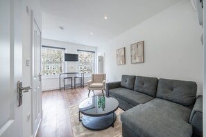 A modern living room with a grey sectional sofa, a round coffee table, and a beige armchair. There are two windows with a view of trees and buildings, and a flat-screen TV mounted on the wall. - Charming One Bedroom Flat in Knightsbridge (London)
