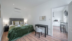 A bedroom with a bed, bedside table, and a desk with a chair. There is a kitchenette in the background. - Charming One Bedroom Flat in Knightsbridge (London)