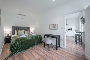 A bedroom with a bed, bedside table, and a desk with a chair. There is a kitchenette in the background. - Charming One Bedroom Flat in Knightsbridge (London)