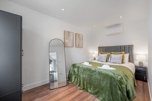 A modern bedroom with a large bed, a mirror, two wall art pieces, a nightstand, and a wardrobe. - Top Floor One-bed With View in Knightsbridge (London)