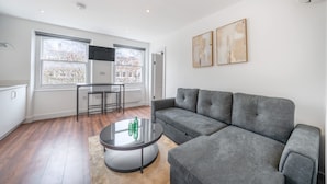 A modern living room with a grey sofa, a round coffee table, and a dining area with a table and chairs. - Top Floor One-bed With View in Knightsbridge (London)
