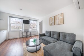 A modern living room with a grey sofa, a round coffee table, and a dining area with a table and chairs. - Top Floor One-bed With View in Knightsbridge (London)