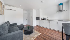 A modern living room with a sofa, a coffee table, and a kitchen area with white cabinets and a stove. - Top Floor One-bed With View in Knightsbridge (London)