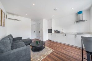 A modern living room with a sofa, a coffee table, and a kitchen area with white cabinets and a stove. - Top Floor One-bed With View in Knightsbridge (London)