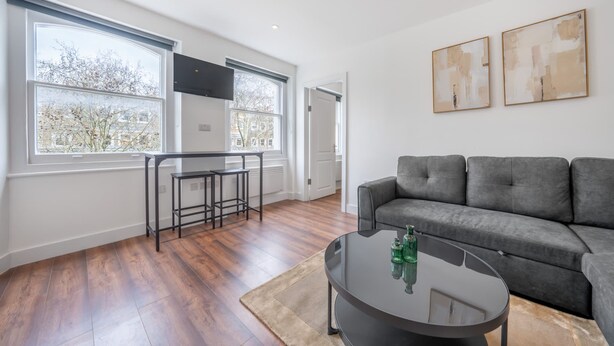 A modern living room with a grey sofa, a round coffee table, and a TV mounted on the wall. - Top Floor One-bed With View in Knightsbridge (London)