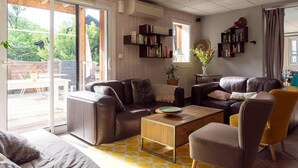 A living room with a sofa, armchair, and coffee table. There are bookshelves and a window with a view of greenery. - Séjour montagne près du domaine skiable | Piscine (Ax-Les-Thermes)