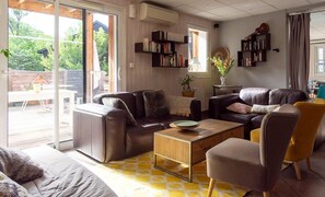 Ein Wohnzimmer mit Sofa, Sessel und Couchtisch. Bücherregale und ein Fenster mit Blick auf Grünflächen.