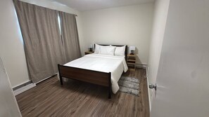 A hotel room with a wooden bed, white bedding, a nightstand with a lamp, a window with curtains, and a door. - Cozy 2-Bedroom Apartment Near Downtown Calgary (Calgary)