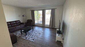 A living room with a brown leather sofa, a black coffee table, a patterned rug, a white TV stand, and a floor lamp. - Cozy 2-Bedroom Apartment Near Downtown Calgary (Calgary)