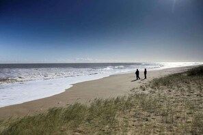 Uma praia com duas pessoas caminhando pela orla, ondas quebrando na areia e um céu limpo.
