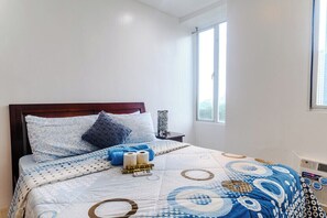 A neatly made bed with a patterned bedspread, a bedside table with a lamp, and a window with a view of buildings. - City Comfort at Seibu Tower (Taguig City)