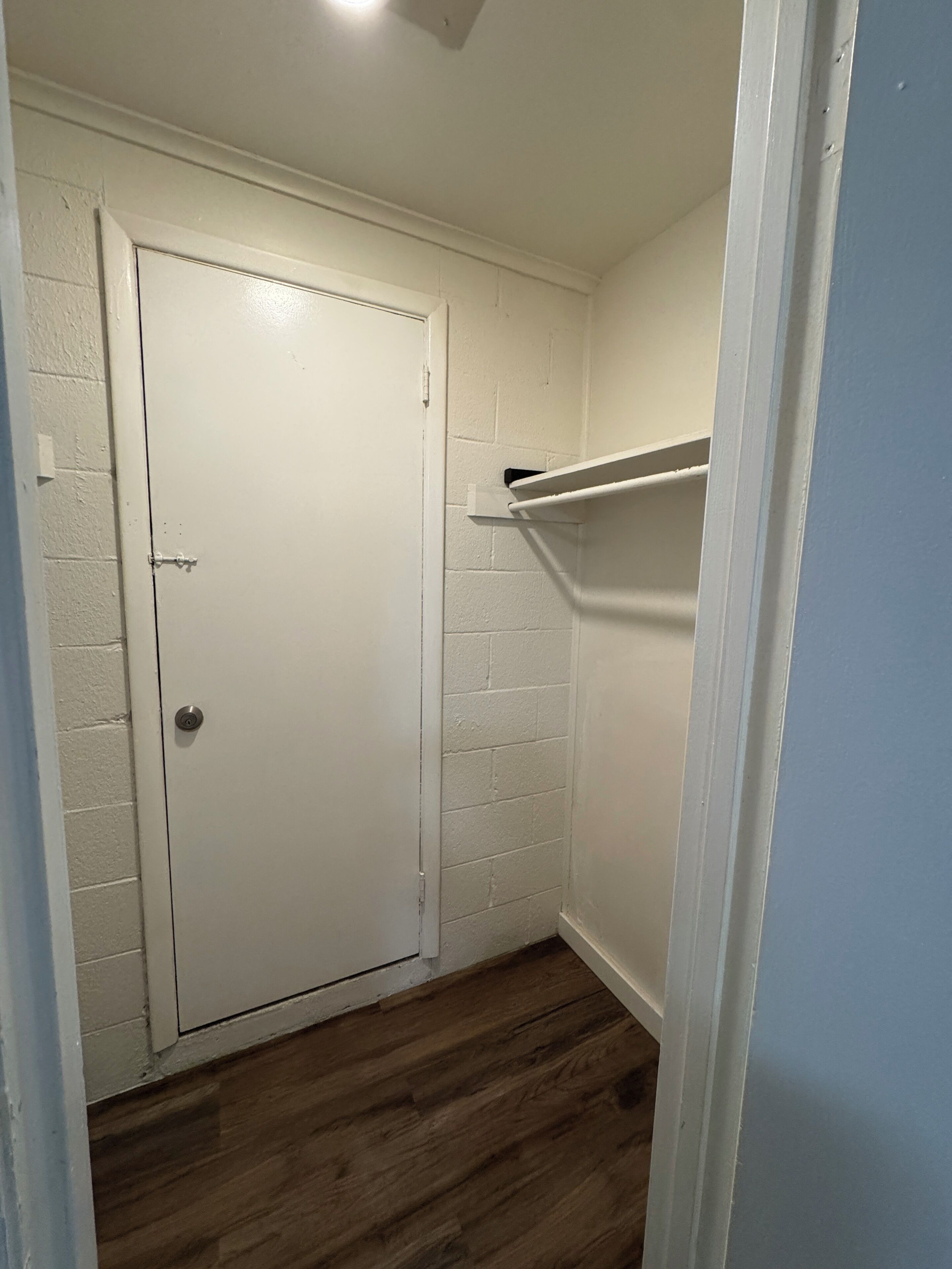 A narrow hallway with a white door, a wooden floor, and a white shelf against the wall.