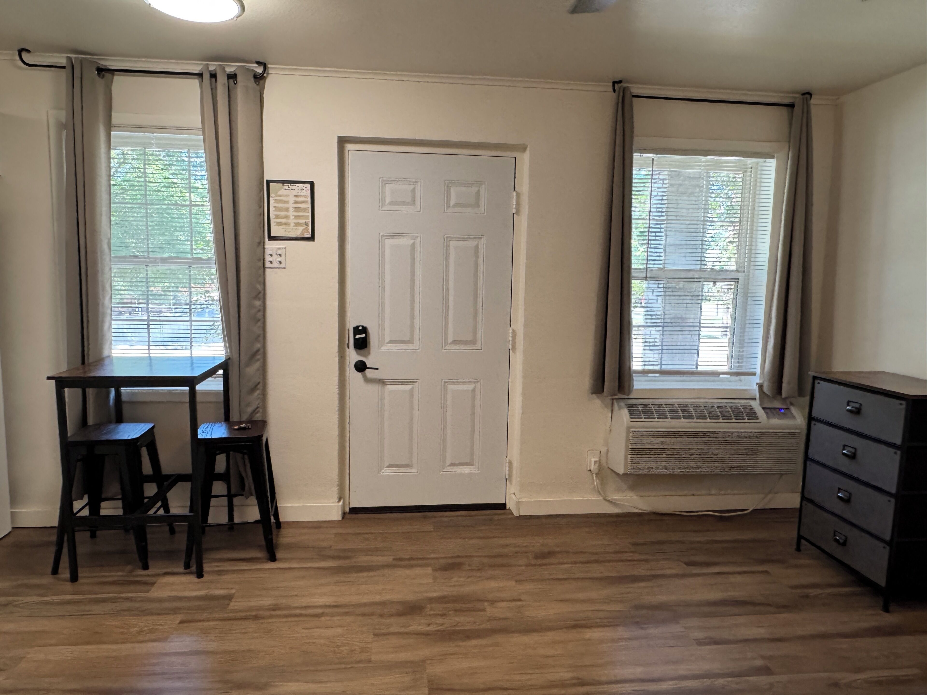 A room with a white door, two windows with blinds, a wooden table with two chairs, a radiator, and a dresser.