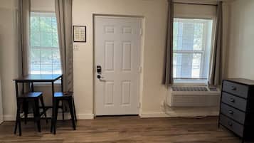 A room with a white door, two windows with blinds, a wooden table with two chairs, a radiator, and a dresser.