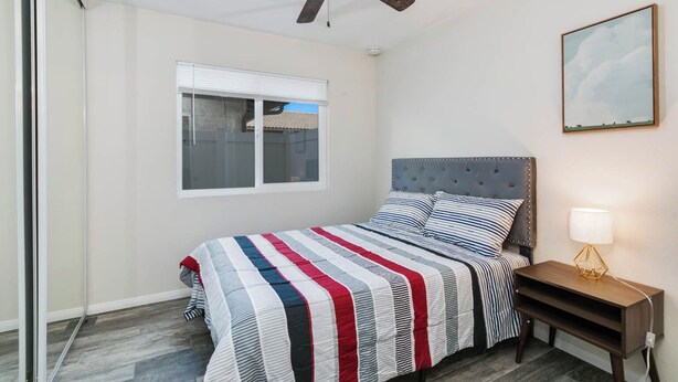 Ein Schlafzimmer mit einem Bett, einem Deckenventilator, einem Fenster, einem Nachttisch mit Lampe und einem gerahmten Bild an der Wand.