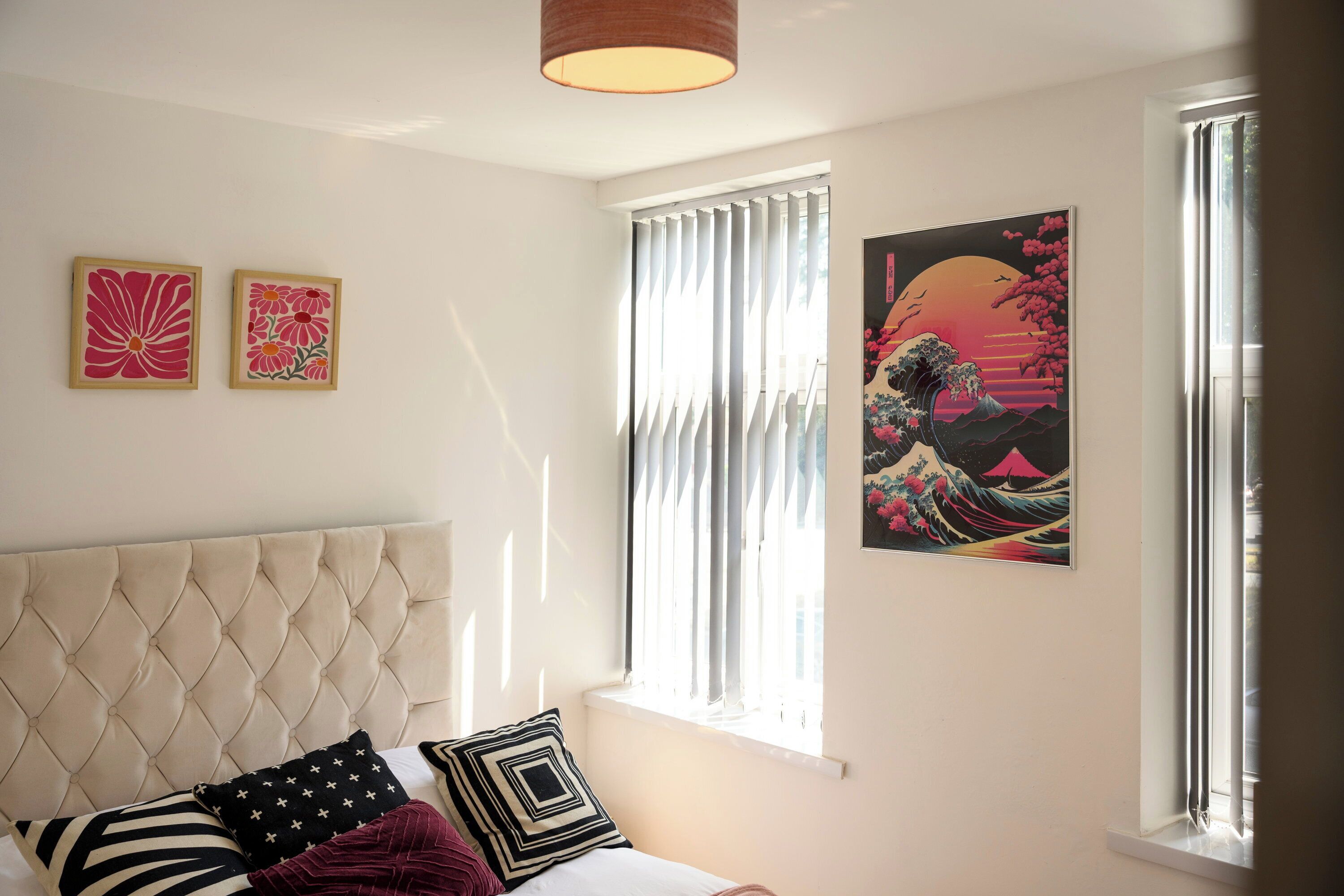 Ein Schlafzimmer mit einem gesteppten Kopfteil, einem Bett mit vielen Kissen, einem Fenster mit Jalousien und zwei gerahmten Kunstwerken an der Wand.