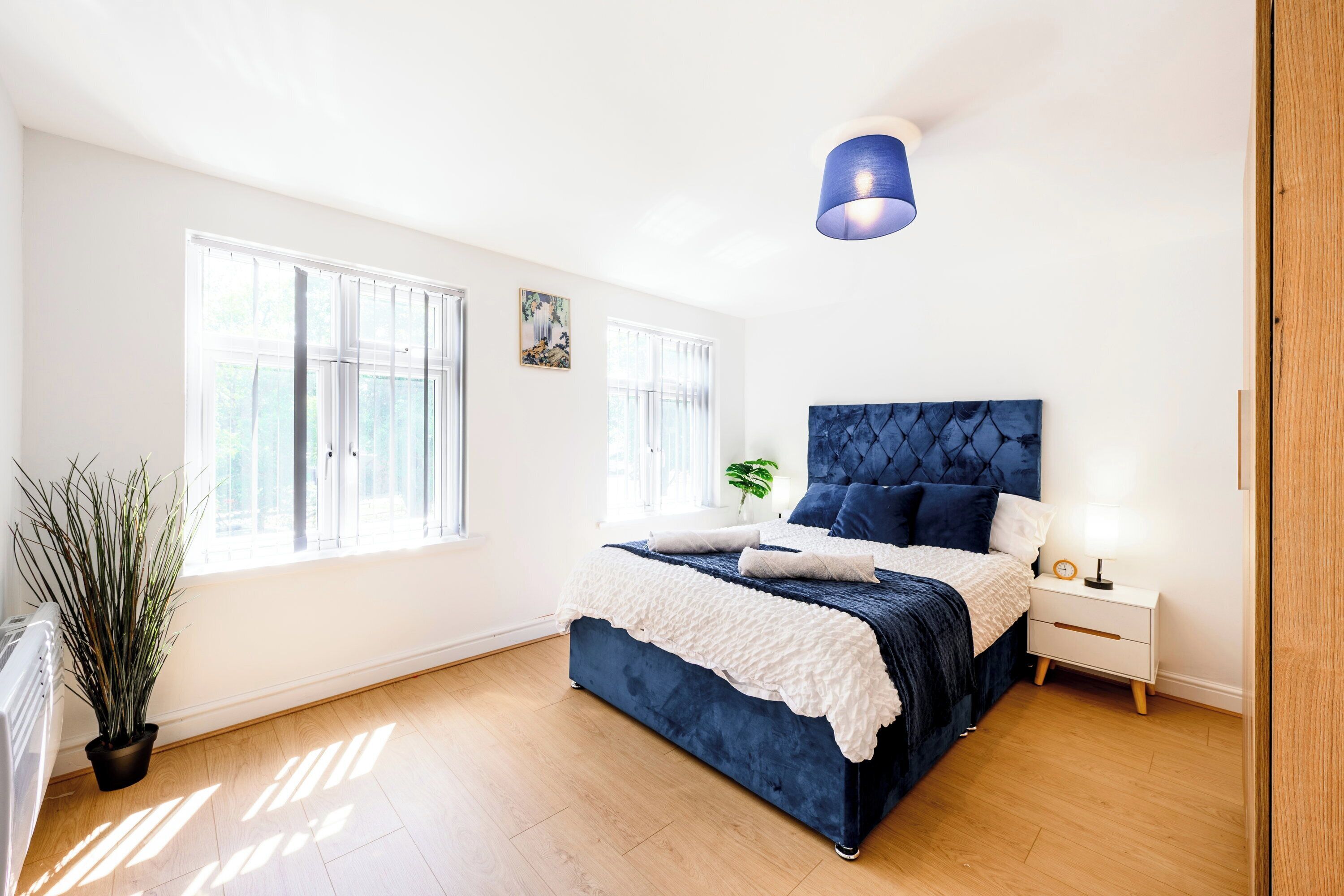 A bedroom with a large bed, a bedside table, a nightstand, a potted plant, and a window.