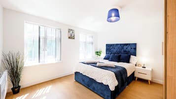 A bedroom with a large bed, a bedside table, a nightstand, a potted plant, and a window.