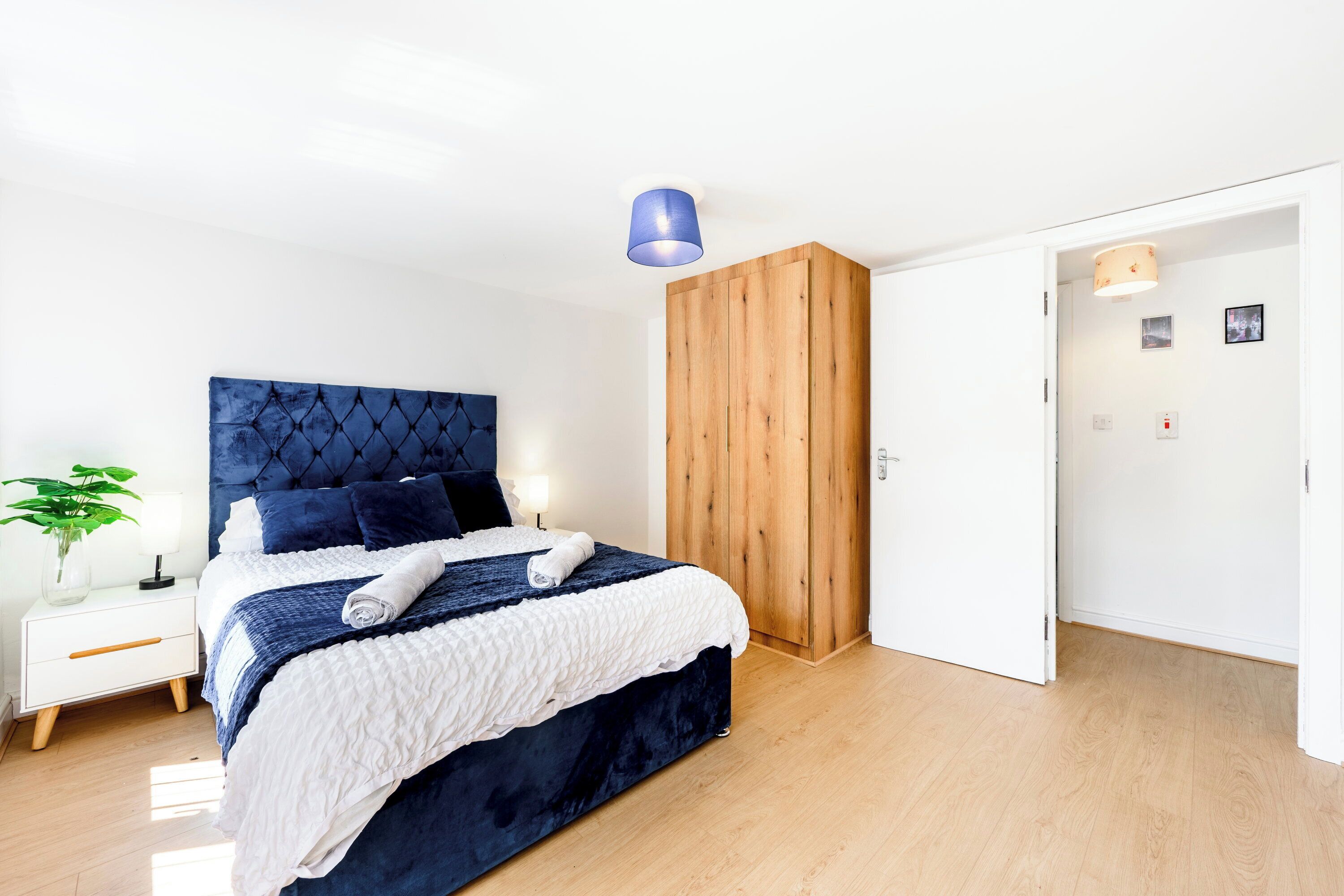 Une chambre à coucher moderne avec une tête de lit capitonnée, une armoire en bois, une table de chevet avec une lampe et une plante en pot.